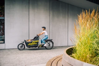 Eine Frau fährt mit dem Ca Go FS200 Life an einem Gebäude vorbei. Die Transportbox ist mit dem Magura IBS Logo versehen.