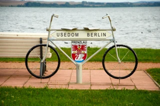 Ein Schild des Radweges in Form eines Fahrrads mit der großen Aufschrift "Usedom Berlin". Darunter ist das Prenzlauer Wappen abgebildet. Im Hintergrund ist ein Fluss.