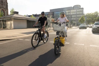 Eine Frau und ein Mann fahren mit Fahrrädern über eine von Autos befahrene Straße.