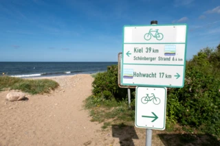 Ein Strandweg führt zur strahlend blauen Ostsee. Im Vordergrund steht ein Schild, welches den Radweg anzeigt.