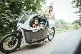 Eine Frau fährt mit dem FS200 Life Family Plus über einen Feldweg. In der Transportbox sitzen zwei Kinder.