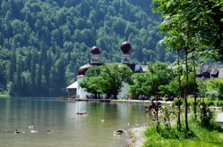 Ein grün-blauer See umgeben von Bäumen. Im Hintergrund ist ein kleines Schloss.