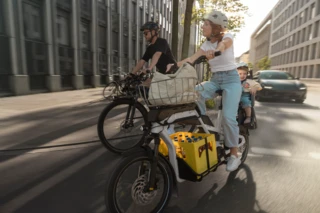 Eine Frau fährt mit dem CS durch ie Stadt. Auf dem hinteren Gepäckträger ist ein Kindersitz montiert, in dem ein kleines Kind sitzt. Daneben fährt ein Mann mit einem Fahrrad.